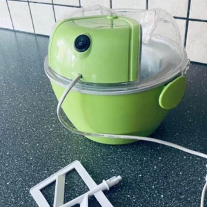 Glassmaskin - Grön elektrisk glassmaskin med transparent lock och avtagbara delar. Utrustad med enkel strömbrytare och medföljande vispdel. Maskinen ser ren och välvårdad ut, utan synligt slitage eller defekter. Perfekt för hemmagjord glass och desserter.