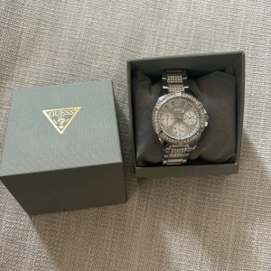 Guess silverfärgad klocka med stenar - Säljer en riktigt snygg och glittrig klocka från Guess i silverfärgat metallmaterial. Urtavlan är dekorerad med stenar och har tre mindre visartavlor. Armbandet är också prytt med stenar och har en klassisk länkdesign. Perfekt för dig som gillar bling och stil.