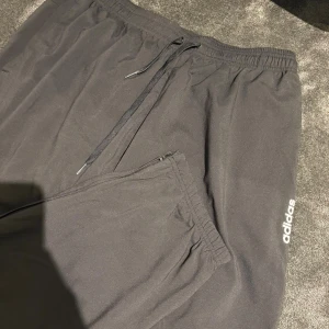 Svarta mjukisbyxor från Adidas XL - Svarta mjukisbyxor från Adidas i storlek XL. Byxorna har resår i midjan med snörning och dragkedjeförsedda fickor. Adidas-logga i vitt på benet. Perfekta för chill eller träning, tillverkade i mjukt syntetmaterial.