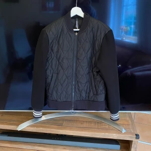 Svart quiltad bomberjacka från Neil Barrett - Snygg svart bomberjacka från Neil Barrett med quiltat mönster på fram- och baksidan. Jackan har svarta ärmar, ribbade muddar med vita ränder och hel dragkedja framtill. Perfekt för dig som vill ha en stilren look