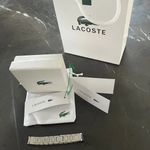 Silverfärgad armbandsklocka från Lacoste - Stilren armbandsklocka från Lacoste i silverfärgat metallarmband med länkdesign och ingraverad Lacoste-logga. Klockan levereras i originalförpackning med tillhörande påse och etiketter. Perfekt accessoar för dig som gillar klassisk och sportig stil. Nypris 1300.