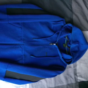 Blå hoodie från BOSS med dragkedja - Snygg blå hoodie från BOSS med hel dragkedja och huva. Tröjan har svarta detaljer på ärmarna och en stilren design. Perfekt för dig som gillar modern stil. Mjuk och skön, passar till många outfits.