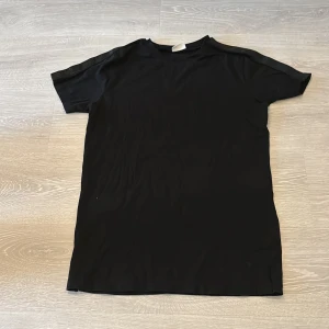 Svart t-shirt från Purewhite - Svart t-shirt från Purewhite i stilren design. T-shirten har korta ärmar och ett diskret band med logga längs axlarna. Perfekt för dig som gillar en clean och enkel look. Materialet känns mjukt och följsamt mot kroppen.