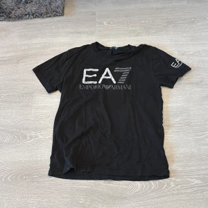 Svart EA7 Emporio Armani t-shirt XL - Svart t-shirt från EA7 Emporio Armani med stort silverfärgat logotryck på bröstet och mindre tryck på ärmen. Klassisk rund hals och korta ärmar. Tillverkad i mjuk bomull för en skön känsla. Perfekt för dig som gillar stilrena och exklusiva plagg.
