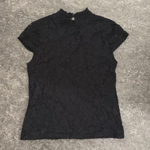 Svart spetsblus med hög krage - Svart blus i spets med blommigt mönster och hög krage. Korta ärmar och knapp i nacken för en extra detalj. Perfekt för dig som gillar en stilren men ändå romantisk look.