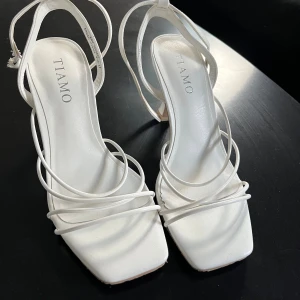 Vita sandaler med klack från Tiamo - Snygga vita sandaler från Tiamo med smala remmar och fyrkantig tå. Skorna har en elegant hög klack och justerbar rem runt vristen. Tillverkade i syntetmaterial med stilren design som passar perfekt till sommarens outfits. De är 7cm klack och använda en gång vilket var på balen🤍