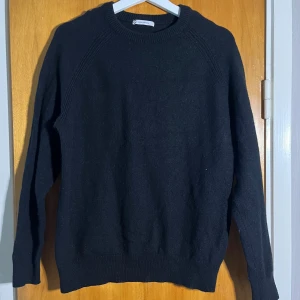 Svart stickad tröja i bomull/ull - En svart stickad tröja storlek L. Tröjan är tillverkad i en mjuk bomulls- och ullblandning, har rund halsringning och långa ärmar. Perfekt för lager-på-lager under kyliga dagar.