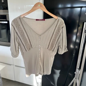 Blus med spets från Indiska - Supersöt blus från Indiska med v-ringning, knappar framtill och fina spetsdetaljer på axlarna. Blusen har trekvartsärm med knapp vid ärmslut. Storlek M ❤️Färgen är väldigt svår att uppfatta men beige/lite åt de gröna hållet?😂