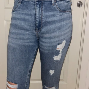 Hollister high-rise croppade jeans - Säljer ett par ljusblå high-rise jeans från Hollister med croppad längd och slitna detaljer på benen. Jeansen har klassisk femficksdesign, skinny fit och är tillverkade i stretchigt denim för en bekväm passform. Storlek xs/s 