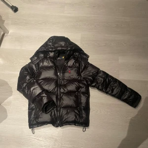 Svart pufferjacka från Polo Ralph Lauren - Säljer en svart pufferjacka från Polo Ralph Lauren med luva och dragkedja. Jackan har en röd broderad logga på bröstet och justerbara ärmslut med kardborre. Perfekt för alla kalla dagar på vintern. Riktigt snygg jacka!🍾