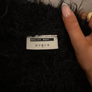 Svart fluffig tröja från Noisy May - Mysig svart fluffig tröja från Noisy May med lång ärm och relaxed fit. Perfekt för kalla dagar när du vill vara både bekväm och snygg. Tröjan har en enkel rund halsringning och är supermjuk med sin lurviga textur.