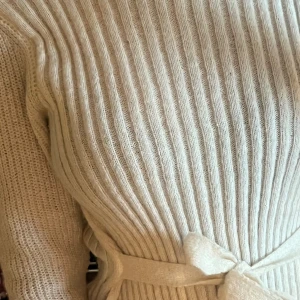 Beige ribbad stickad tröja med bälte(knuten kan fixas) - Supermysig beige stickad tröja med ribbat mönster och knytbälte i midjan. Långärmad och figurnära passform som ger en snygg siluett. Perfekt för dig som vill ha en stilren och bekväm look.