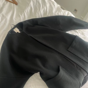 Svart lång kappa utan krage och med fickor  - Snygg svart lång kappa med raka linjer och stora framfickor. Jackan har minimalistisk design utan krage och knyts i midjan med bälte. Perfekt för dig som gillar stilrena och trendiga plagg. Passar till många olika outfits.