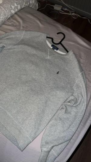Grå sweatshirt från Polo Ralph Lauren - Snygg grå sweatshirt från Polo Ralph Lauren med klassisk rund hals och diskret blå logga broderad på bröstet. Tröjan har långa ärmar och ribbade muddar vid ärmslut och nederkant. Perfekt för chill dagar och enkel att matcha med jeans eller joggers.