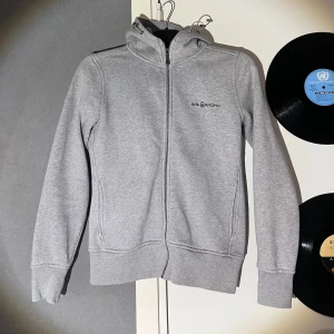 Grå hoodie från Sail Racing - Grå hoodie från Sail Racing med dragkedja hela vägen och broderad logga på bröstet. Tröjan har huva, långa ärmar och två sidofickor. Materialet är mjuk bomullsmix med insida i fleece för extra komfort. Perfekt för chill dagar och snygg till jeans.