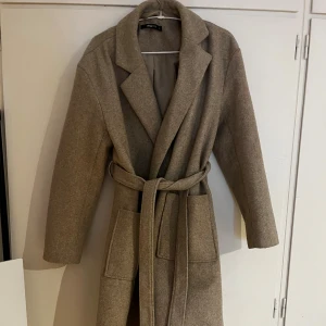 Beige kappa från Gina Tricot med bälte - Säljer en stilren beige kappa från Gina Tricot i storlek 34. Den har klassisk krage, stora fickor framtill och ett matchande bälte i midjan. Materialet är mjukt och känns som ullblandning, perfekt för höst och vinter. Enkel och snygg att slänga på sig när det är kallt ute.