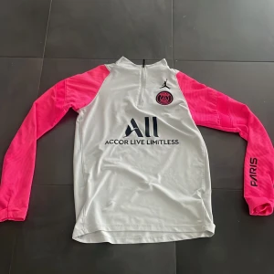 PSG x Jordan half zip träningströja - Snygg PSG x Jordan träningströja i ljus grå med starkt rosa ärmar och svart logga. Half zip framtill, långärmad och med broderad Paris Saint-Germain emblem på bröstet. Perfekt för fotboll eller sport, tillverkad i mjuk polyester. Färgerna passar bra ihop och sitter slim.