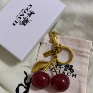 Coach nyckelring med glittriga körsbär - Superfin nyckelring från Coach med två glittriga körsbär i rött och detaljer i guldfärgad metall. Perfekt accessoar till väskan eller nycklarna, med Coach-logga på både ring och tag. Kommer med original ask och tygpåse.