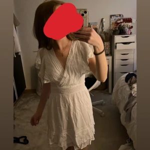 Vit omlottklänning med volang - Supersöt vit kortklänning med omlottliv, v-ringning och volangdetaljer vid ärmar och kjol. Klänningen har smock i midjan som ger en snygg passform och är tillverkad i ett lätt, luftigt material. Perfekt för varma dagar och somriga vibes! Aldrig använd! 