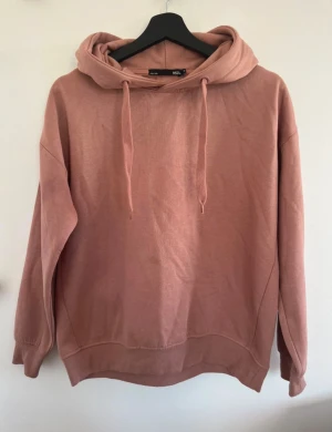 Rosa hoodie från 157 med huva - Rosa hoodie från 157 med stor huva och snörning framtill. Tröjan har långärmade armar och är gjord i mjuk bomull. Passformen är loose och den har ribbade muddar vid ärmslut och nederkant. Perfekt för sköna dagar och häng.