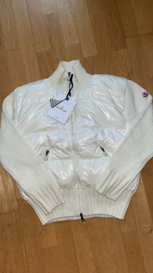 Vit pufferjacka från Moncler - Exklusiv vit pufferjacka från Moncler med stickade ärmar och krage. Jackan har glansig yta på kroppen, två fickor med dragkedja och Moncler-logga på vänster ärm. Materialmix av polyester och stickad ull ger en unik look. Perfekt för dig som vill ha något stilrent och lyxigt.