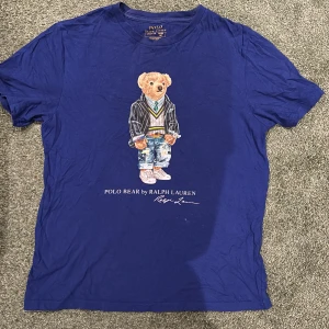 Blå Polo Bear t-shirt från Ralph Lauren - Säljer en blå t-shirt från Ralph Lauren med ikoniska Polo Bear-trycket på bröstet. T-shirten har rund hals och korta ärmar. Nypris 600. Skick 8/10