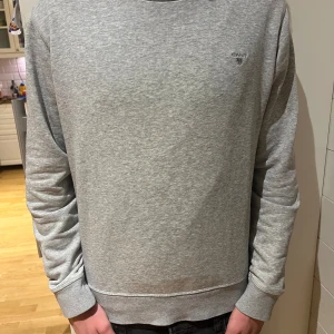 Grå sweatshirt från GANT - Snygg grå sweatshirt från GANT med diskret logga på bröstet. Tröjan har rund hals, långa ärmar och ribbade muddar vid ärmslut och nederkant. Perfekt för chill dagar och enkel att matcha med jeans eller joggers.