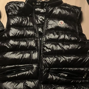 Svart dunväst från Moncler - Svart, glansig dunväst från Moncler med klassisk logga på bröstet. Västen har quiltad design, två fickor med dragkedja och hög krage. Perfekt för lager-på-lager och har en snygg, normal passform. Stängs med dragkedja framtill.