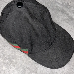Svart keps från Gucci med monogram - Säljer en svart keps från Gucci med diskret GG-monogram över hela kepsen. Klassisk modell med böjd skärm och ikoniska gröna och röda ränder på sidan. Perfekt accessoar för att lyfta din streetwear-look.