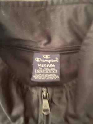 Svart vindjacka från Champion M - Svart vindjacka från Champion i storlek medium. Jackan har dragkedja framtill, höga krage och vita logoband längs ärmarna med Champion-tryck. Tillverkad i polyester, perfekt för sportiga dagar. Snygg och enkel design med fickor på sidorna.