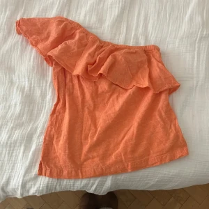 Orange offshoulder topp med volang - Säljer en snygg orange offshoulder topp med bred volang längs halsringningen. Toppen har en asymmetrisk design med ena axeln bar och är tillverkad i ett lätt linnematerial. Perfekt för dig som gillar färg och vill sticka ut lite extra.