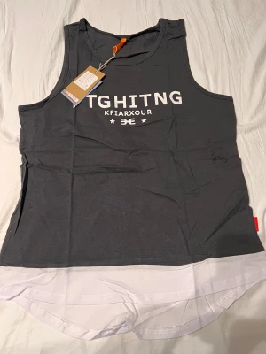 Grå och vit linnetopp med RUN-tryck - Säljer en cool grå linnetopp från KFIARXOUR med vit text framtill och stort RUN-tryck på ryggen. Toppen har rund hals och ärmlös design, med vit kontrastkant nedtill. Perfekt för dig som gillar sportig stil och vill sticka ut på träningen.