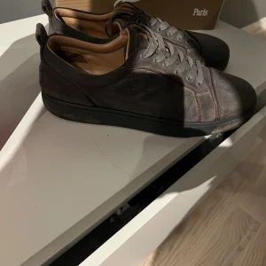 Christian Louboutin grå sneakers mocka - Snygga svarta sneakers från Christian Louboutin i mocka med klassisk röd sula. Skorna har rund tå, platt sula och grå snörning. Insidan är beige och de har en stilren, minimalistisk design som passar till många outfits.