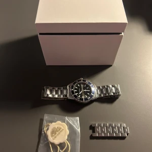 Leijona silverfärgad armbandsklocka - Stilren armbandsklocka från Leijona med rund svart urtavla och silverfärgat metallarmband. Klockan har tydliga markeringar, datumvisning och blå bezel. Extra länkar och originalbox medföljer. Nypris 800 kr. barn storlek men var allt för stor för min handled, därför tog jag av massor med länkar. Klocka passar därmed även vuxna. Men ett ganska litet klock ur. 