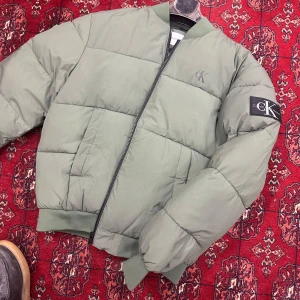 Grön pufferjacka från Calvin Klein Jeans - Snygg grön pufferjacka från Calvin Klein Jeans med svart innerfoder och dragkedja framtill. Jackan har ribbade muddar, diskret CK-logga på bröstet och patch på ärmen. Tillverkad i återvunnen polyester och nylon. Perfekt för kyliga dagar.obs kolla på bild 4 och 5 lite skada men inget förhinder annars helt ny 