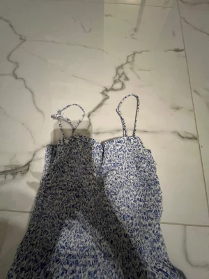 Blommig långklänning med smock - Säljer en söt långklänning med blått och vitt blommigt mönster. Klänningen har smock upptill och tunna axelband, som också är justerbara . Materialet känns lätt och luftigt, perfekt för varma dagar. Passformen är normal och klänningen har en lite flowy siluett.