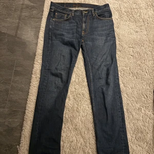 Mörkblå jeans från Nudie Jeans 30/32 - Snygga mörkblå jeans från Nudie Jeans i klassisk straight fit. Jeansen har fem fickor, orangea kontrastsömmar och en stilren design. Tillverkade i slitstarkt denim med normal passform och medelmidja. 