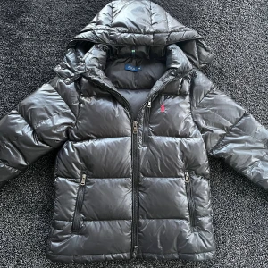 Svart dunjacka från Polo Ralph Lauren - Snygg svart dunjacka från Polo Ralph Lauren med huva och två dragkedjeförsedda fickor. Jackan har en quiltad design och klassisk röd Polo-logga på bröstet. Skick 9/10, Storlek M, OBS! du vet varför den är billig.                         Pris går att diskutera✅