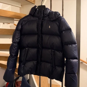 Marinblå dunjacka från Polo Ralph Lauren - Snygg, populär och slutsåld marinblå dunjacka från Polo Ralph Lauren med huva, två fickor med dragkedja och klassisk logga på bröstet. Jackan har puffad design, hög krage och stängs med både dragkedja och tryckknappar. Perfekt för kalla dagar. Köpt förra året och är sparsamt använd. En defekt (se bild 4) tappat en knapp går säkert att fixa. Tar självklart bort sminket på kragen (se bild 4) innan jag postar. Köpt för ca 4000. TAR INTE EMOT SKAMBUD.