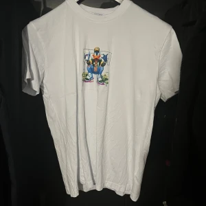 Vit Off White t-shirt med tryck - Vit t-shirt från Off White med färgglatt tryck av en tecknad anka i streetwear-stil på både fram- och baksidan. Stort 'OFF WHITE'-tryck på ryggen. Skön passform och klassisk rund hals. Perfekt för dig som gillar unika och lekfulla motiv.