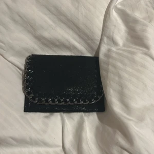 Svart plånbok med kedjedetalj - Svart plånbok i skinn med glansig finish och silverfärgad kedja runt kanten. Plånboken har en fyrkantig form och stängs med en magnetknapp. Perfekt storlek för kort och kontanter. 💞