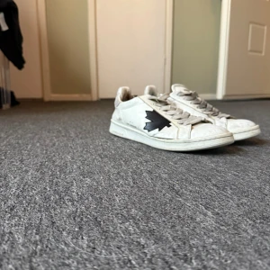 Vita sneakers från Dsquared2 - Säljer ett par vita sneakers från Dsquared2 med svart lönnlöv på sidan. Skorna har snörning, rund tå och platt sula. Tillverkade i skinn med Dsquared2-logga bak på hälen. Perfekta för dig som gillar stilrena och trendiga sneakers.