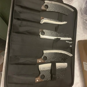 Handsmidd köksknivset med förvaringsväska - Exklusivt set med 5 handsmidda köksknivar i robust förvaringsväska. Knivarna har hamrad finish, ergonomiska trähandtag och hål för bättre grepp. Bladen är i nyskick utan synligt slitage eller defekter. Perfekt för både hemmakockar och proffs.