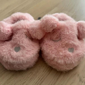 Rosa fluffiga tofflor med nalleansikte Tofflor Storlek 21/22 - Supergulliga rosa tofflor med mjukt fluffigt material och nalleansikte framtill. Broderade ögon och hjärta på nosen, samt små öron som sticker ut. Perfekt för att hålla fötterna varma hemma. Sula med gummidetaljer för extra grepp.Söta rosa tofflor med djurmotiv. Perfekt för små fötter.