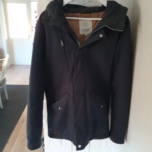 Svart vindjacka från Elvine Cornell - Mörk marinblå/svartaktig vindjacka från Elvine, modell Cornell, med huva och dragkedja framtill. Jackan har två snedställda fickor med tryckknappar och är helfodrad i beige. Materialet är en mix av bomull och polyester, vilket gör den både slitstark och bekväm. Perfekt för kyliga dagar och en clean look. 