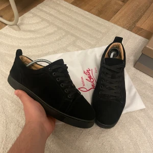 Svarta sneakers från Christian Louboutin - Säljer ett par svarta sneakers från Christian Louboutin med klassisk röd sula och snygg mocka på ovansidan. Skorna har rund tå, platt sula och svart snörning. Insidan är i beige skinn för extra komfort. Perfekt för dig som gillar exklusiva och stilrena sneakers.storlek 41 passar 42  box finns