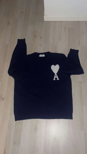 Mörkblå stickad tröja från Ami - Mörkblå stickad tröja från Ami med vit hjärtapplikation och bokstaven A på bröstet. Rund hals, ribbade muddar och nederkant. Perfekt för dig som gillar stilrena detaljer och vill ha något som sticker ut lite extra.