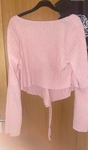 Ljusrosa ribbad långärmad topp - Säljer en ljusrosa ribbad topp med långa ärmar och snygg snörning framtill som skapar en rosett-effekt. Toppen har croppad passform och är tillverkad i mjukt material med vertikala ränder. Perfekt för dig som gillar söta detaljer och en chill vibe.