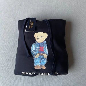 Svart hoodie Polo Bear Ralph Lauren XL - Svart hoodie från Polo Ralph Lauren i storlek XL med ikoniskt Polo Bear-tryck framtill. Tröjan har huva med snörning och ribbade muddar. Materialet är mjuk bomull och polyesterblandning, perfekt för chill dagar. Snyggt tryck med björn i jeans och RL-tröja.