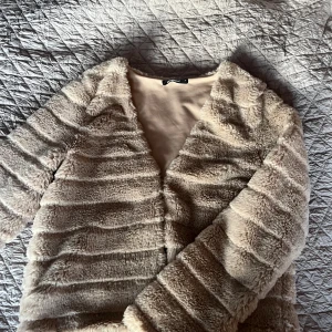 Beige pälsjacka från Gina Tricot - Supermjuk och fluffig beige pälsjacka från Gina Tricot. Jackan har lång ärm, horisontella ränder i pälsen och en öppen front utan synliga knappar eller dragkedja. Perfekt för dig som vill ha en cozy och trendig look under kyliga dagar.